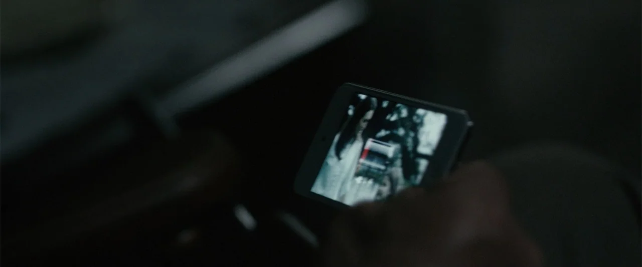 Maggie (2015) movie still: closeup — A dark interior setting shows a person holding a smartphone. The phone screen displays a blurred image of a…