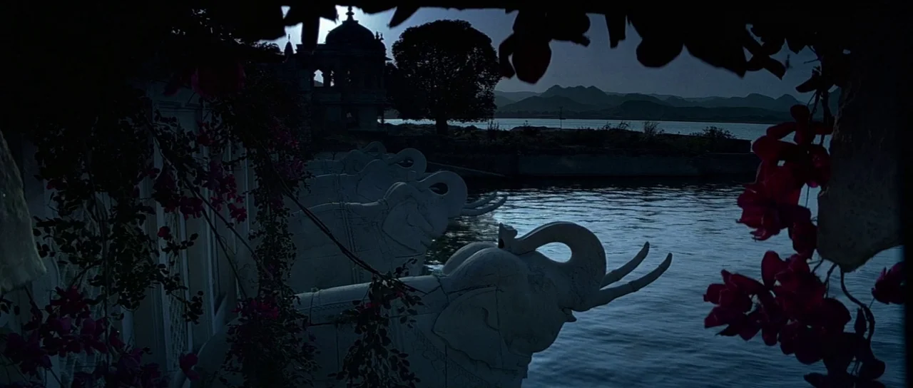 Octopussy (1983) movie still: wide — A row of carved white stone elephant statues overlooks a dark, calm lake. In the distance, silhouettes of hills…
