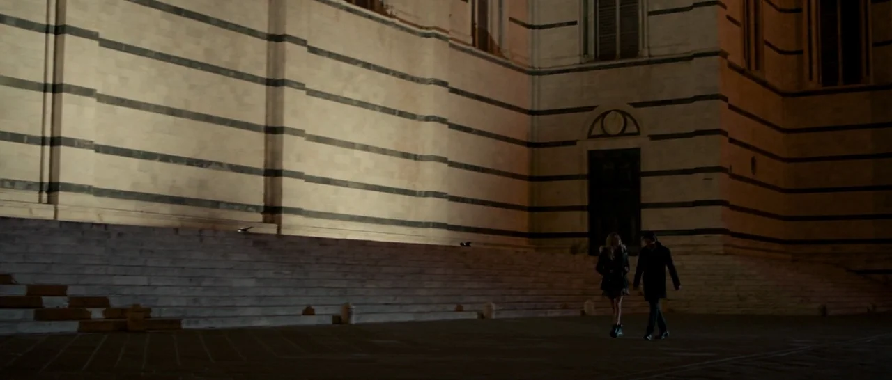 The Face of an Angel (2014) movie still: wide — A man and woman walk across a large, dark, stone-paved plaza at night. A massive building with…