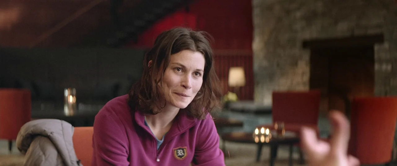 Force Majeure (2014) movie still: medium — A dimly lit hotel lounge area with stone walls and red seating furniture. A woman in a pink jacket…