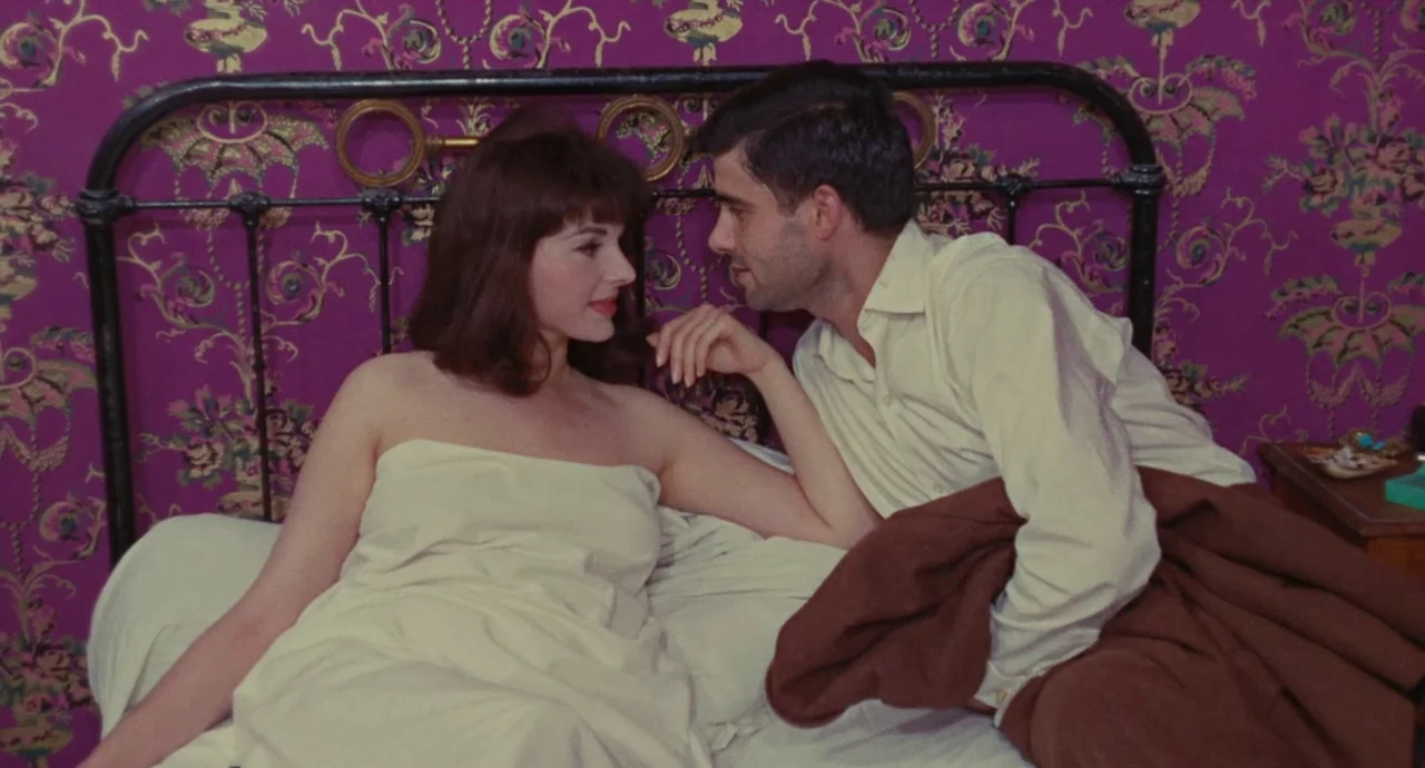 The Umbrellas Of Cherbourg (1964) movie still: medium — A room features bold purple floral-patterned wallpaper and a vintage black metal bed frame.…