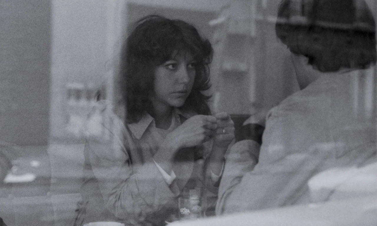 The Secret Son (1979) movie still: medium — A woman and a man sit together at a table, viewed through a window reflection. The woman stares forward…