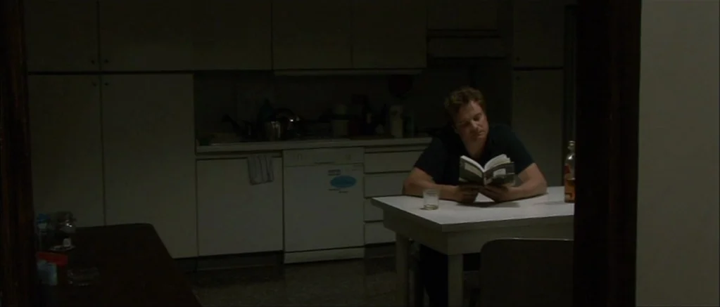 Genova (2008) movie still: medium — A dimly lit room with white cabinetry and a white table. A man sits alone at the table reading a small book.