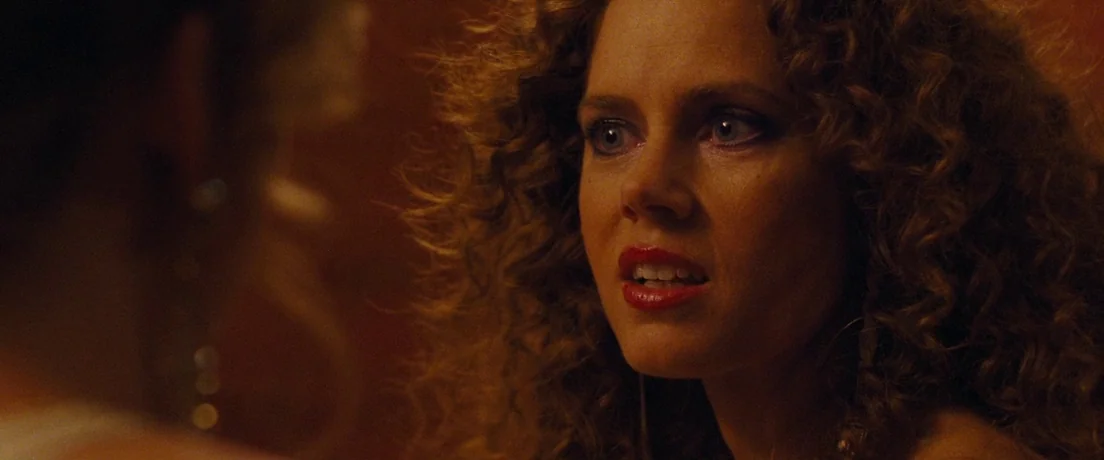 American Hustle (2013) movie still: closeup — A dimly lit room with warm orange tones and shadowed foreground elements. A woman with voluminous…