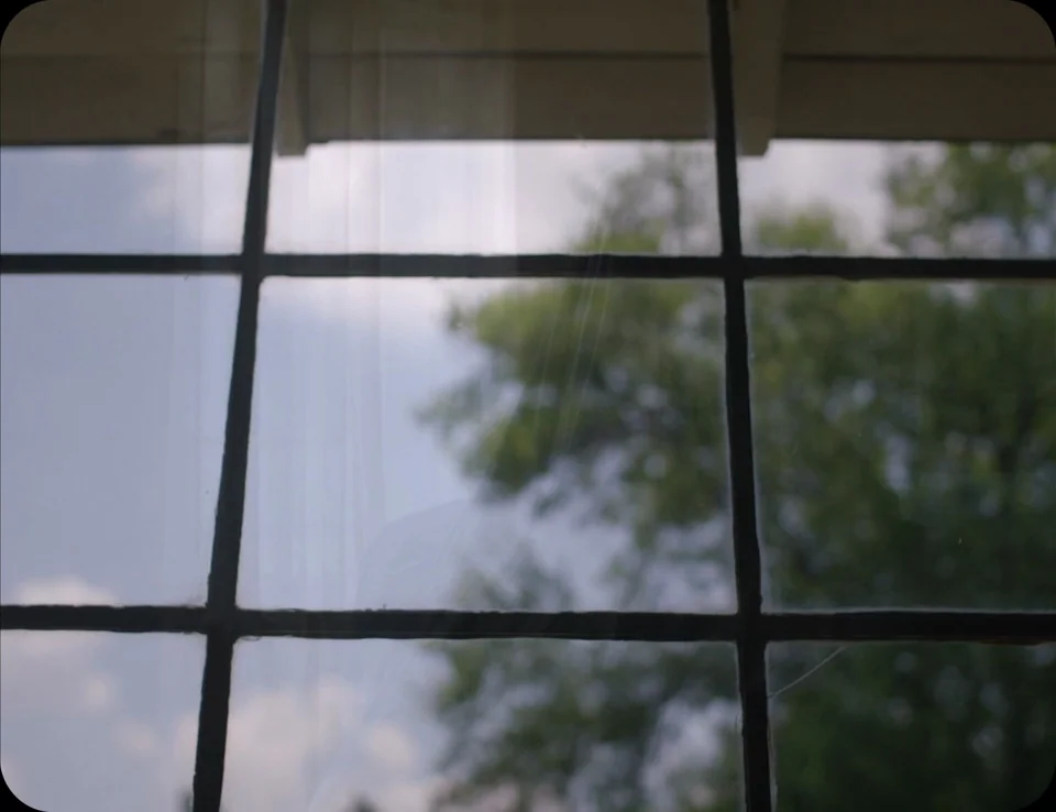 A Ghost Story (2017) movie still: establishing — A multi-pane window with black muntins looks out onto a blurry outdoor scene. Pale sky and green…