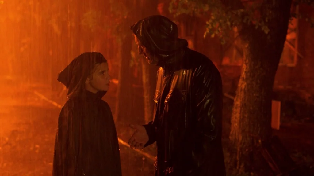 The Ugly Swans (2006) movie still: medium — A dimly lit street at night under heavy rain with orange ambient lighting. A man in a raincoat faces a…