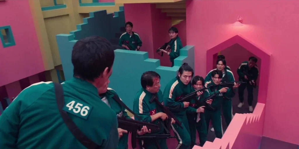 Squid Game (2024) movie still: wide — A surreal M.C. Escher-inspired stairwell features vibrant pink walls and teal stair structures. A group of…