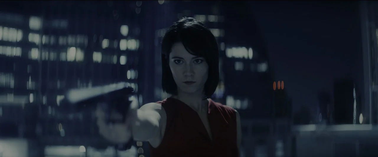 Kate (2021) movie still: medium — A woman stands on a high-rise balcony at night with city lights out of focus. She holds a handgun aimed directly…