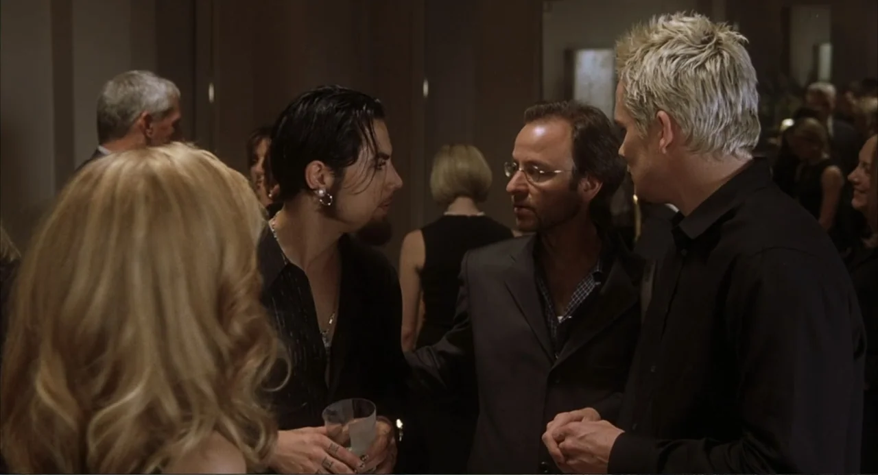Uptown Girls (2003) movie still: medium — A dimly lit indoor social gathering with multiple people in formal evening attire. Three men are huddled…