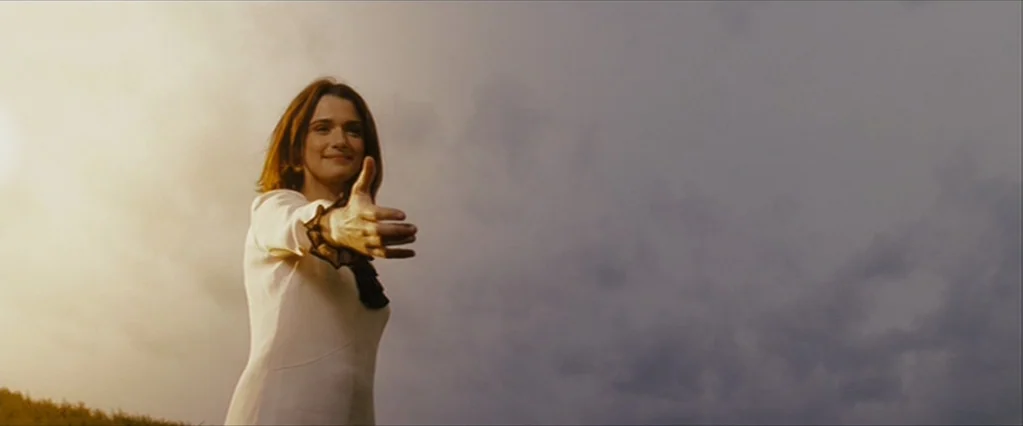 The Brothers Bloom (2008) movie still: medium — A woman stands in a field against a cloudy sky during golden hour. She reaches her arm out toward…