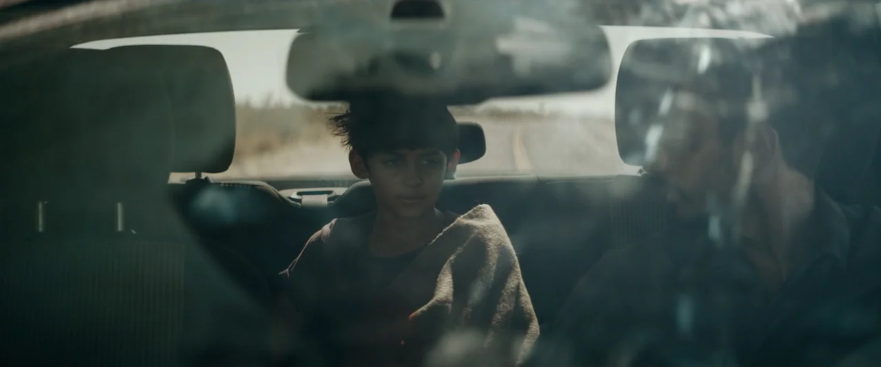 Encounter (2018) movie still: medium — Two individuals sit inside a car cabin during a daylight drive on a road. A young child occupies the backseat…