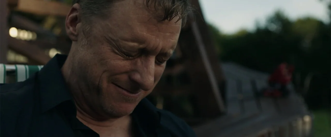 Playing God (2021) movie still: closeup — An outdoor wooden deck area is visible under soft natural late afternoon light. A middle-aged man stands…