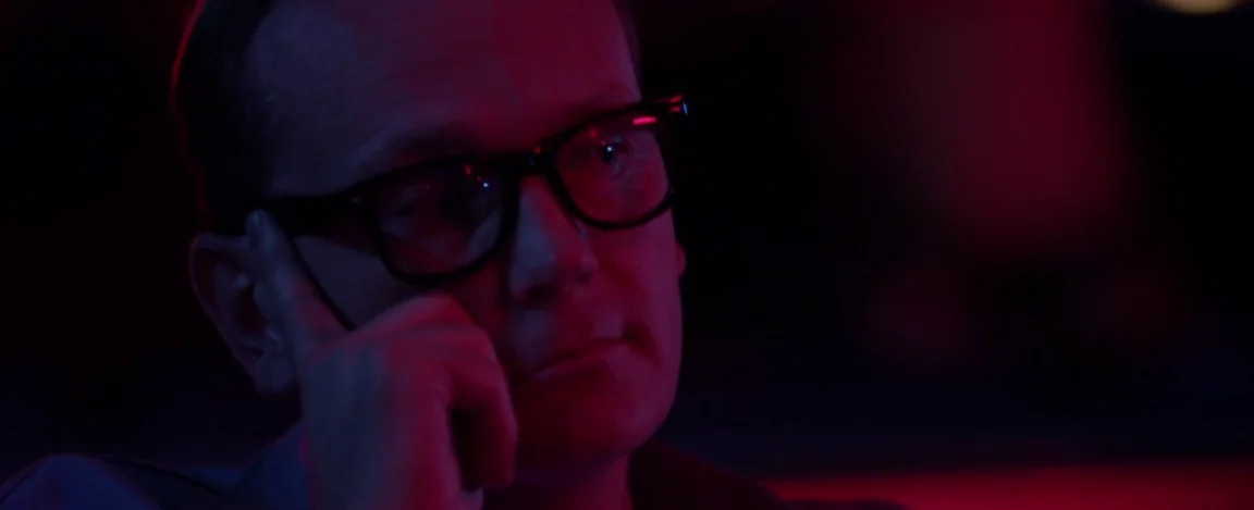 Cheap Thrills (2013) movie still: closeup — A dark, dimly lit space with minimal visible background details. A man sits with his hand against his…
