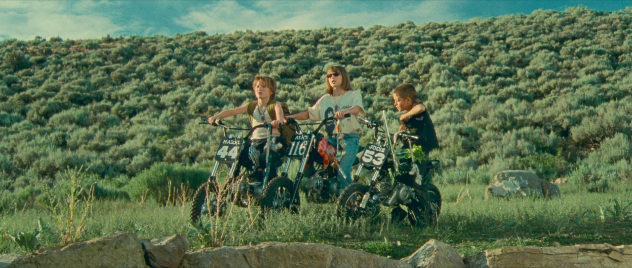 Riddle of Fire (2023) movie still: wide — A grassy, shrub-covered hillside rises in the background under a partly cloudy sky. Three children are…