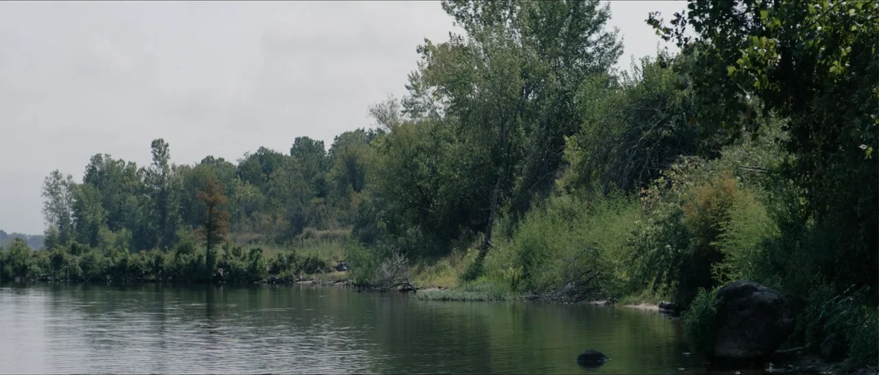 Crescent City (2024) movie still: establishing — A calm river flows alongside a dense line of lush green deciduous trees. The shoreline is filled…