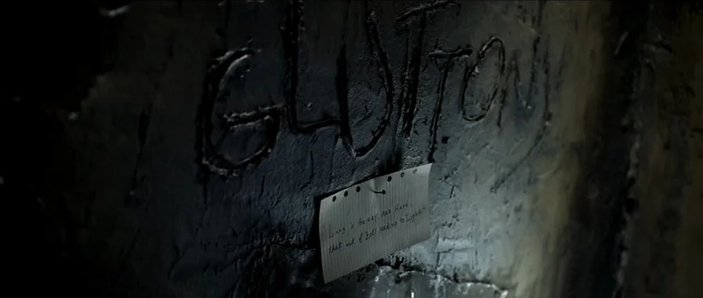 Se7en (1995) movie still: closeup — A dark, uneven surface features the word GLUTTONY carved deeply into the black material. A small, lined piece of…