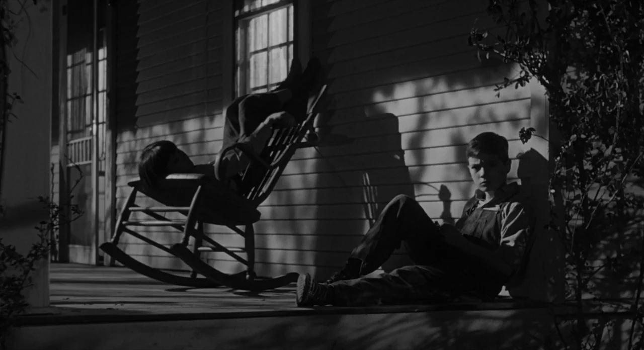 To Kill A Mockingbird (1962) movie still: fullbody — A wooden porch on a clapboard house features sharply defined shadows from sunlight. Two boys…