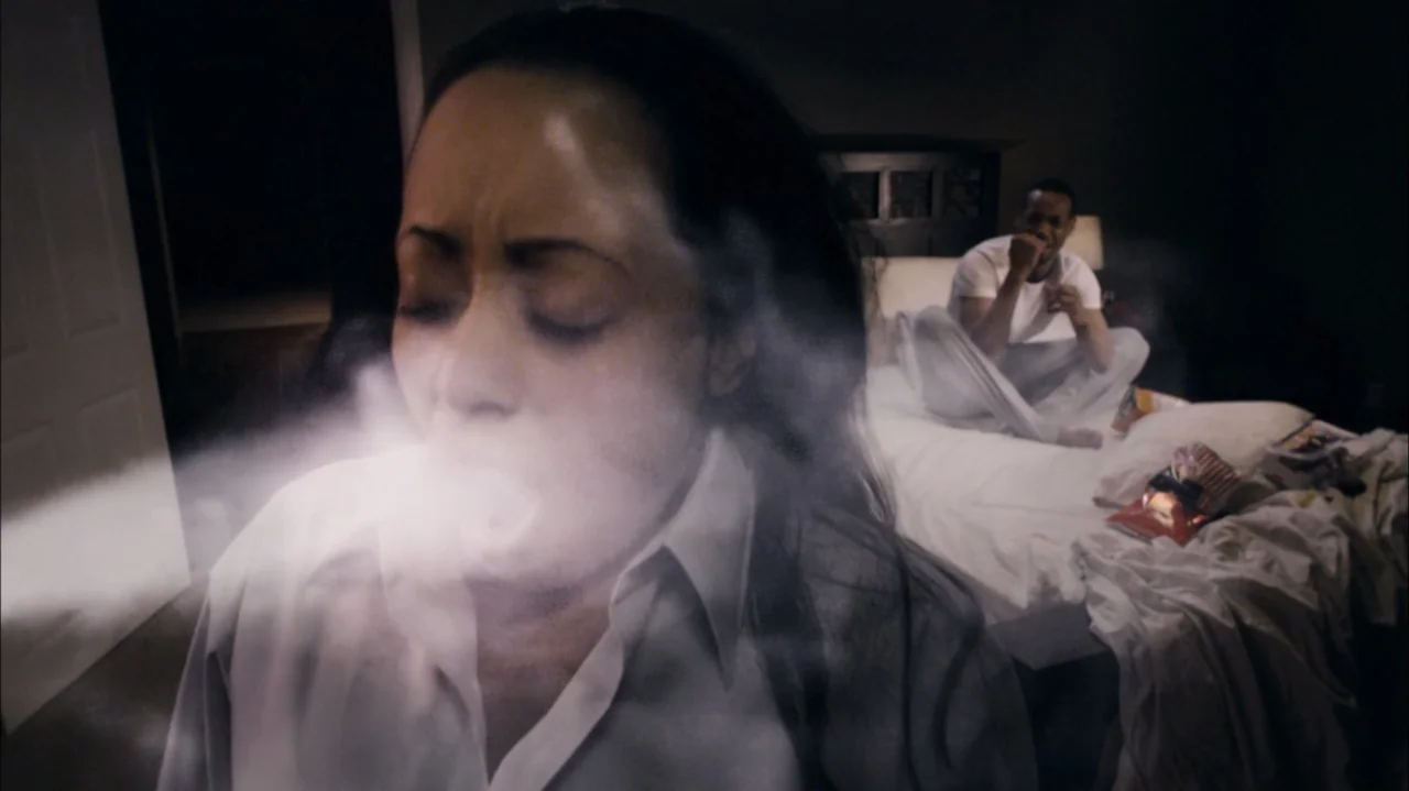 A Haunted House (2013) movie still: closeup — A bedroom at night has a man sitting on a bed in the background. A woman in the foreground exhales a…