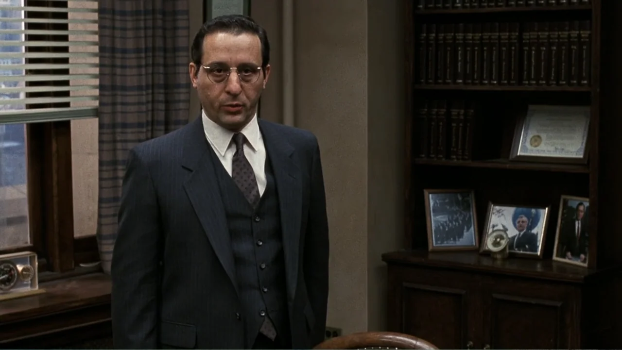 Presumed Innocent (1990) movie still: medium — A professional office with a wooden bookshelf full of binders and framed photographs. A man in a dark…