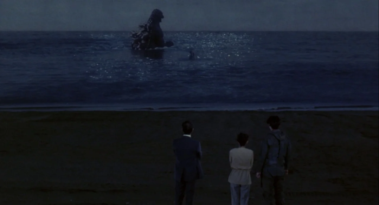 Godzilla vs. Mechagodzilla II (1993) movie still: wide — A dark beach at night meets a turbulent sea reflecting cool moonlight. Three people stand…