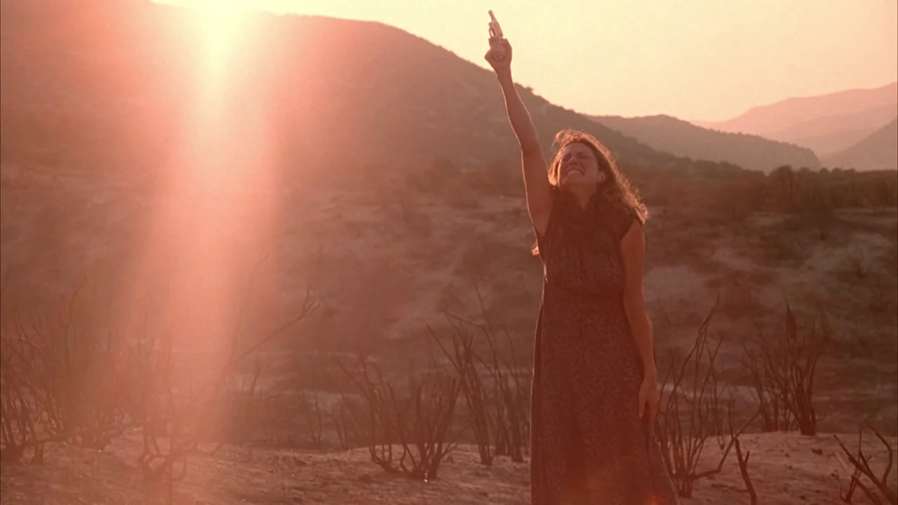 The Rapture (1991) movie still: fullbody — A woman stands in a desolate desert landscape filled with dry, burnt brush. She holds a small pistol…