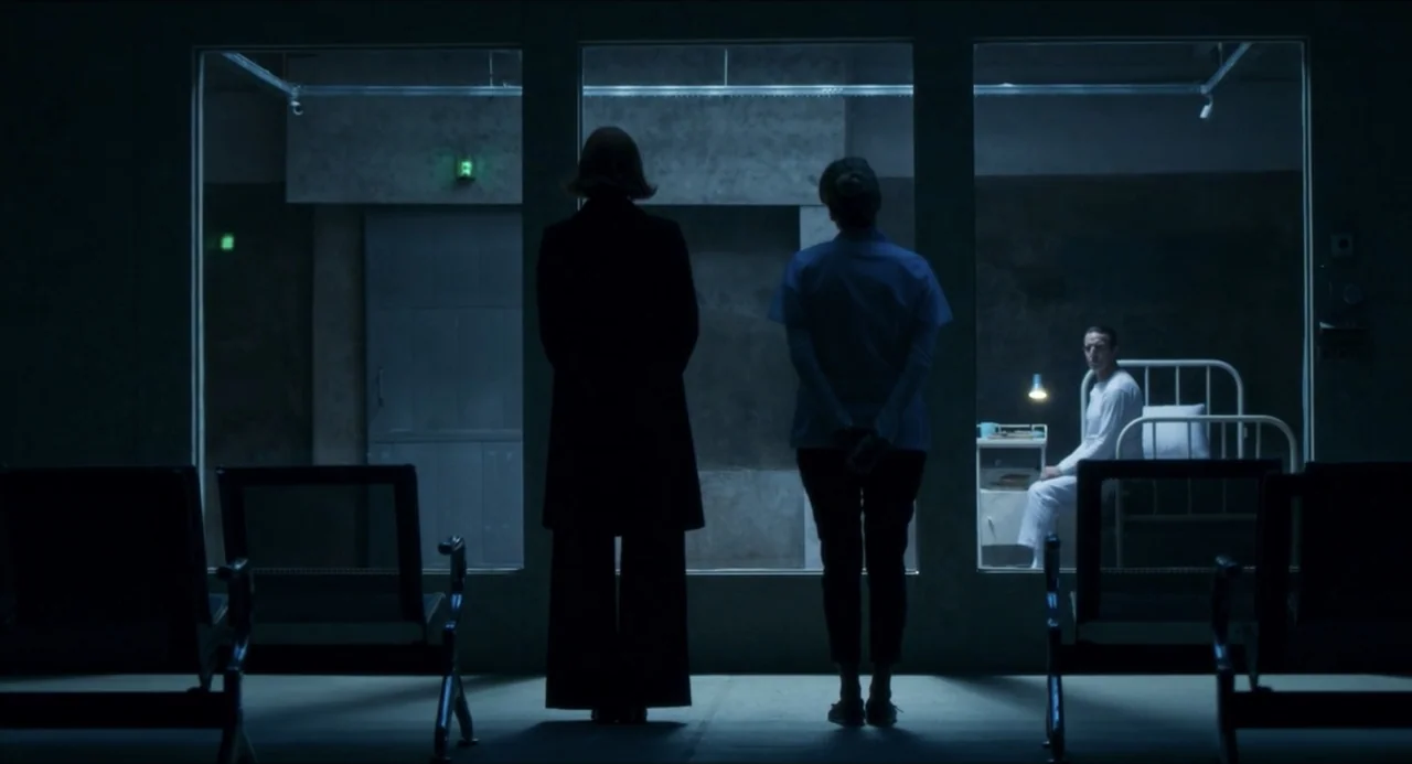 Echoes Of A Crime (2025) movie still: fullbody — A dark waiting area features three large glass panes dividing the space from a clinical unit. Two…