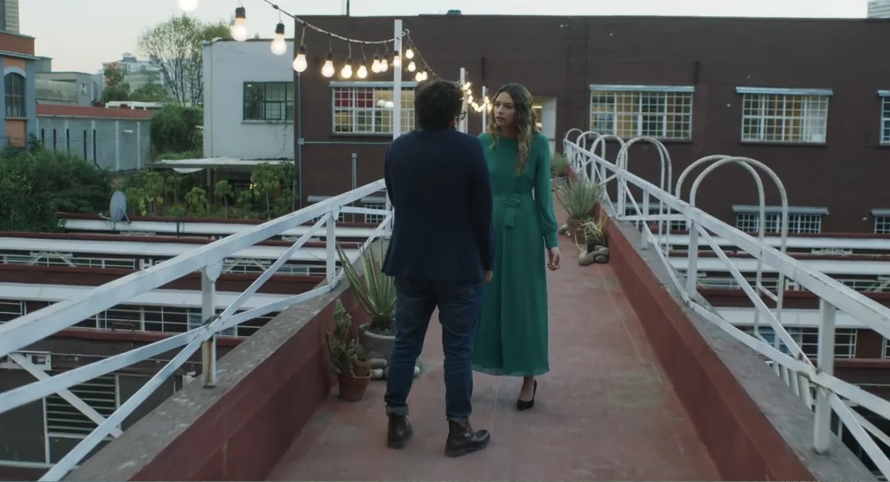 Casando a mi Ex (2023) movie still: fullbody — An outdoor elevated walkway connects two brown brick buildings at golden hour. A man and a woman…