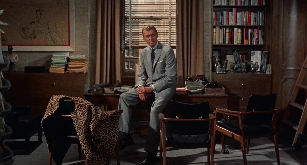 Bell Book and Candle (1958) movie still: medium — A wood-paneled room contains a desk, bookshelves, and various personal items. A man in a suit sits…