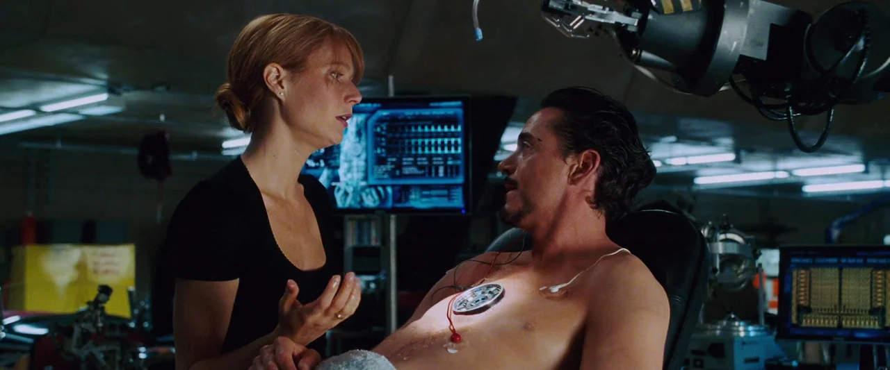 Iron Man (2008) movie still: medium — A dimly lit technological workspace filled with digital monitors displaying data and mechanical arms. A woman…