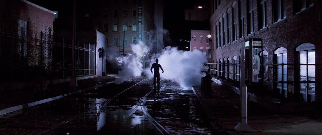 Hackers (1995) movie still: wide — A dark, wet urban street features railroad tracks running down the center pavement. A lone man stands in the…