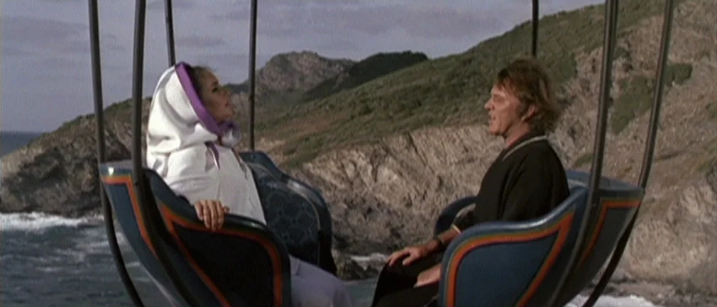 Boom! (1968) movie still: medium — A man and a woman sit in an open gondola lift over a rocky coast. Green hills and the ocean are visible in the…