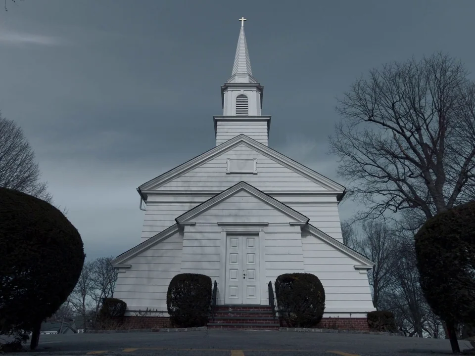 First Reformed (2017) movie still: establishing — A white clapboard church stands against a heavy, grey, overcast sky. The structure is flanked by…