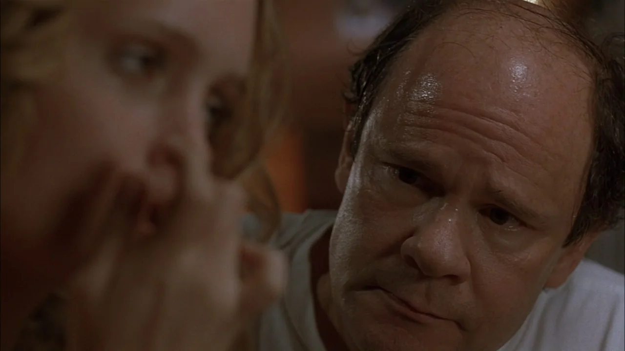 The Babysitters (2008) movie still: closeup — A dimly lit interior room with blurred foreground elements. An older man gazes intently toward an…