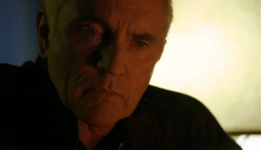 The Limey (1999) movie still: closeup — An interior space lit by the warm glow of an unseen lamp. An older man with short gray hair stares directly…