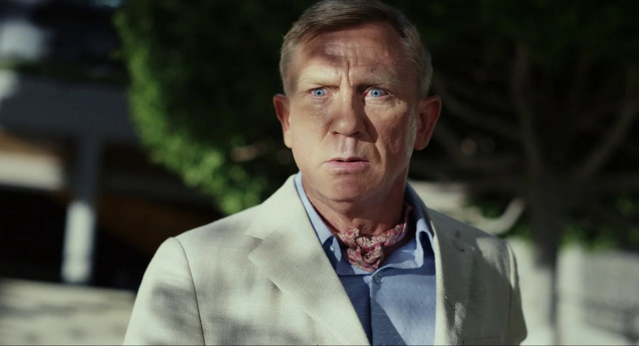 Glass Onion (2022) movie still: closeup — A man stands outdoors in bright daylight near lush greenery and white building structures. He wears a…