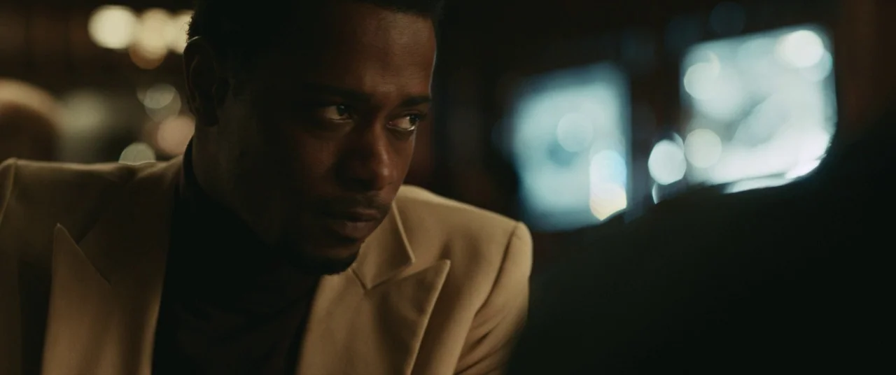 Judas and the Black Messiah (2021) movie still: closeup — A man in a cream blazer sits at a table in a dark, upscale environment. He gazes off-frame…