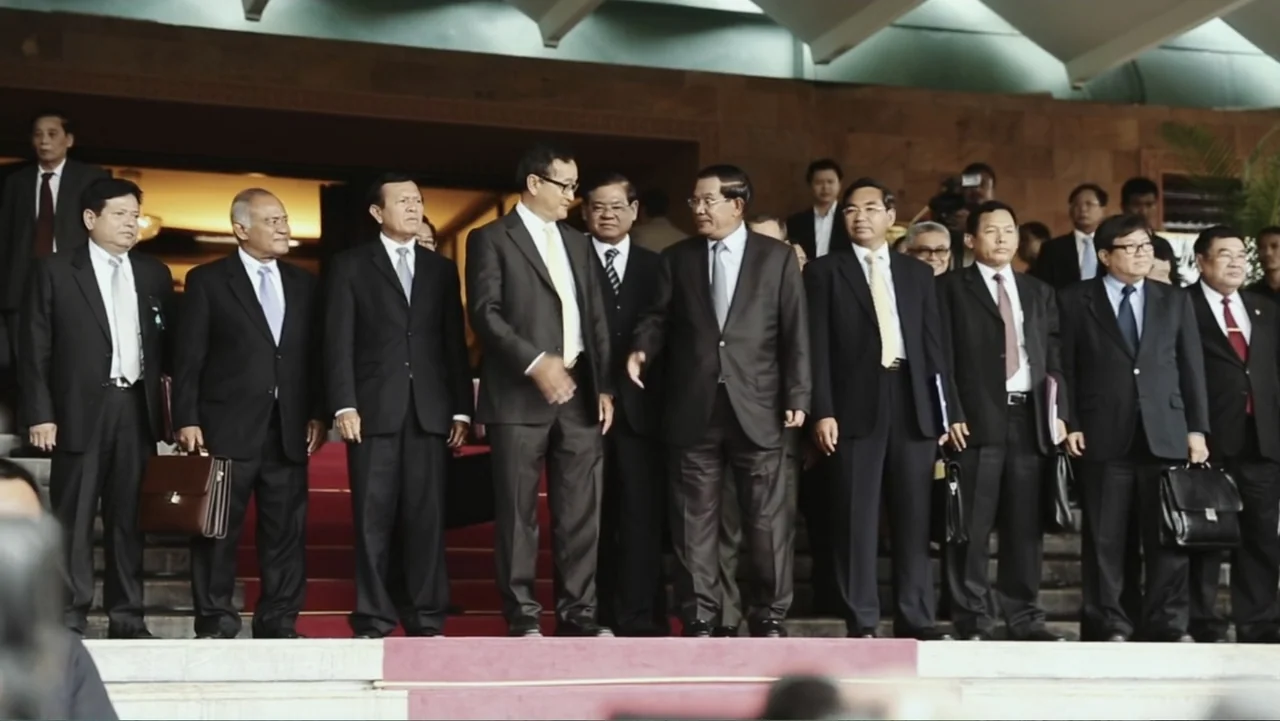 A Cambodian Spring (2018) movie still: fullbody — A line of men in suits stands on the steps of a stone government building. The men are arranged in…