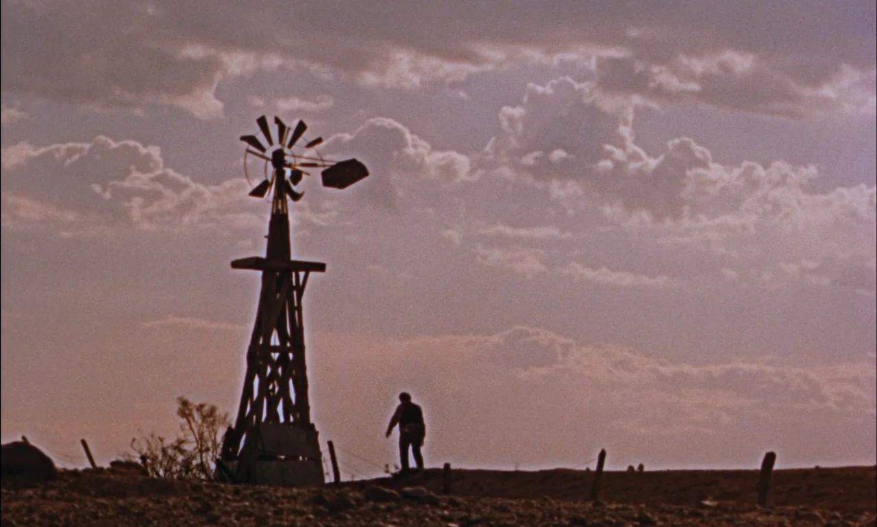 Giant (1956) movie still: wide — A flat, arid field stretches under a cloudy sky during the late afternoon. A man walks in silhouette past a tall,…