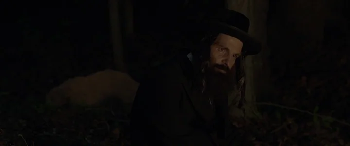To Dust - Nightmares (2018) movie still: medium — A dark, wooded area at night with dense shadows and scattered vegetation. A man wearing a…