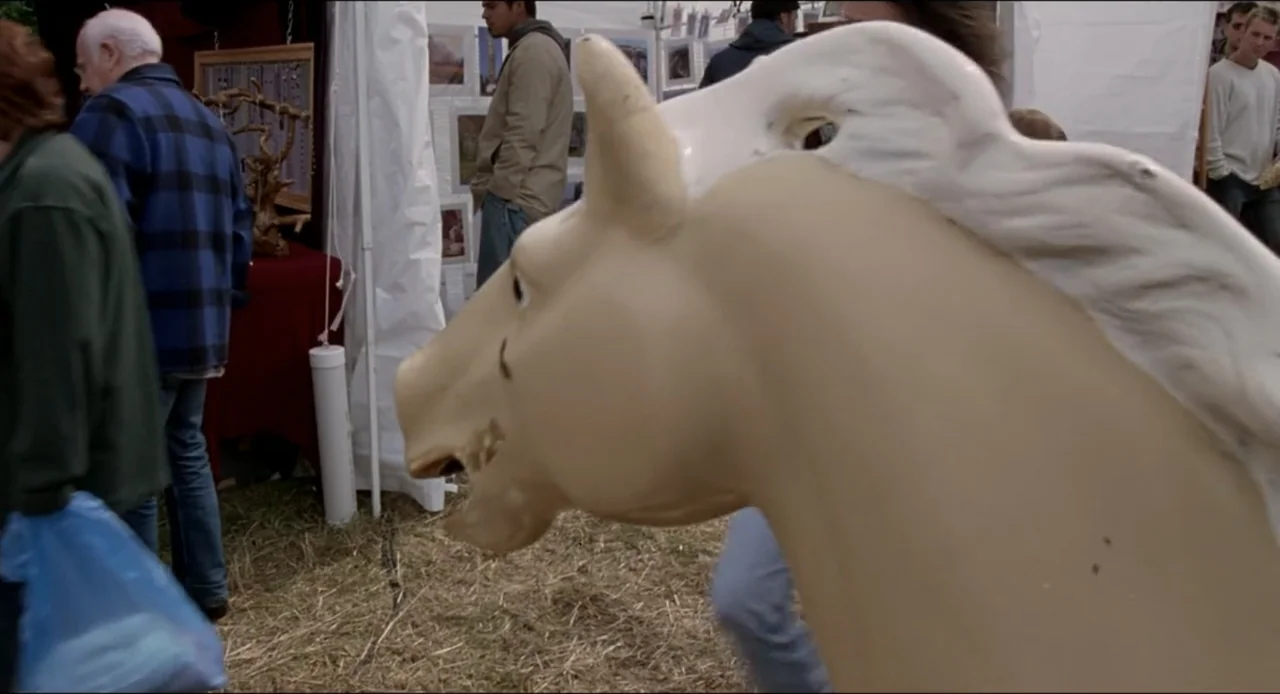 The Ring Two (2005) movie still: closeup — A crowded outdoor craft fair features white vendor tents on a grassy lot. A cream-colored carousel horse…