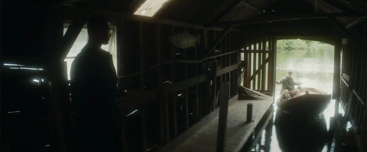 Black Bag (2025) movie still: medium — A wooden boathouse with horizontal wall slats opens onto a calm lake waterway. One man stands inside the…
