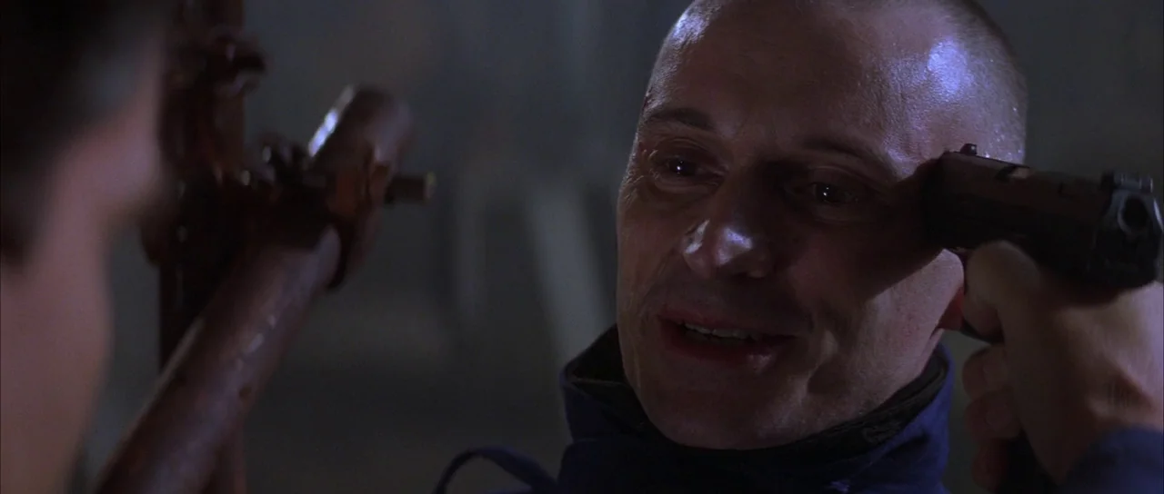 The World is Not Enough (1999) movie still: closeup — A dimly lit industrial setting with indistinct walls behind the man. A man with a shaved head…