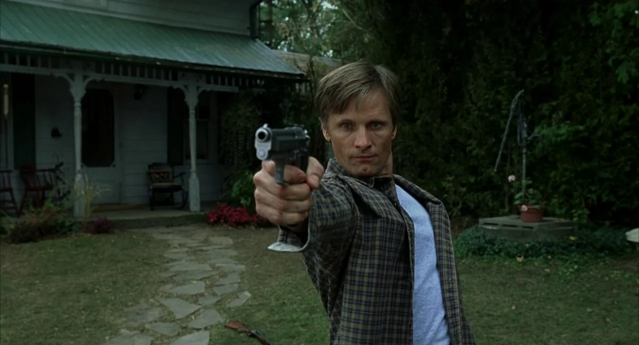 A History of Violence (2005) movie still: medium — A residential front yard features a white house with a green roof porch. A man stands in the…