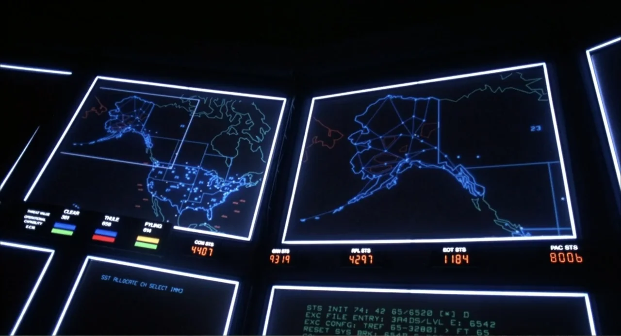 WarGames (1983) movie still: medium — A dark military command center features several large computer displays with glowing map interfaces. These…