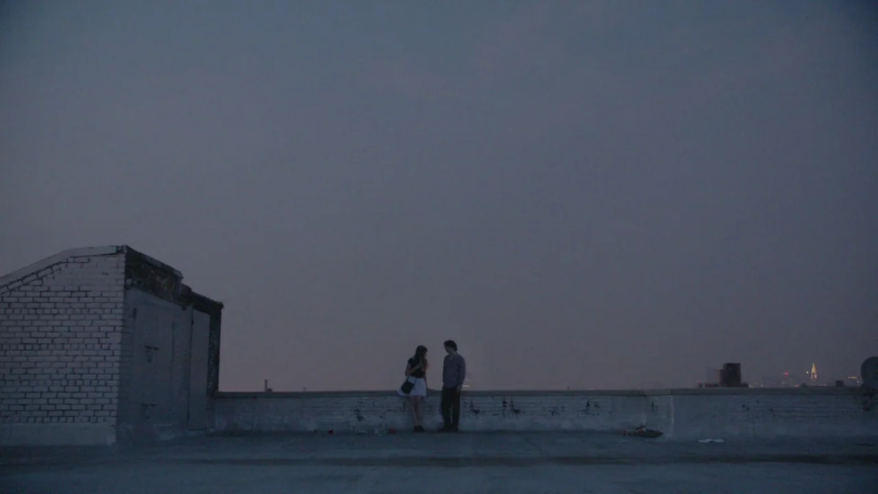 The Heart Machine (2014) movie still: wide — A flat urban roof at dusk with a low white painted brick wall. A young man and a woman stand together…