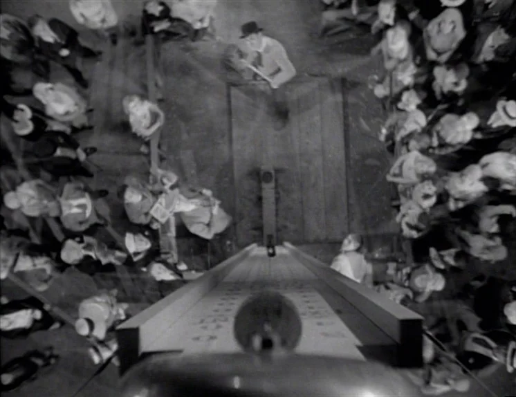 Anna Christie (1930) movie still: wide — A top-down view of a crowd gathered around a high striker carnival game. People in 1930s clothing stand in…