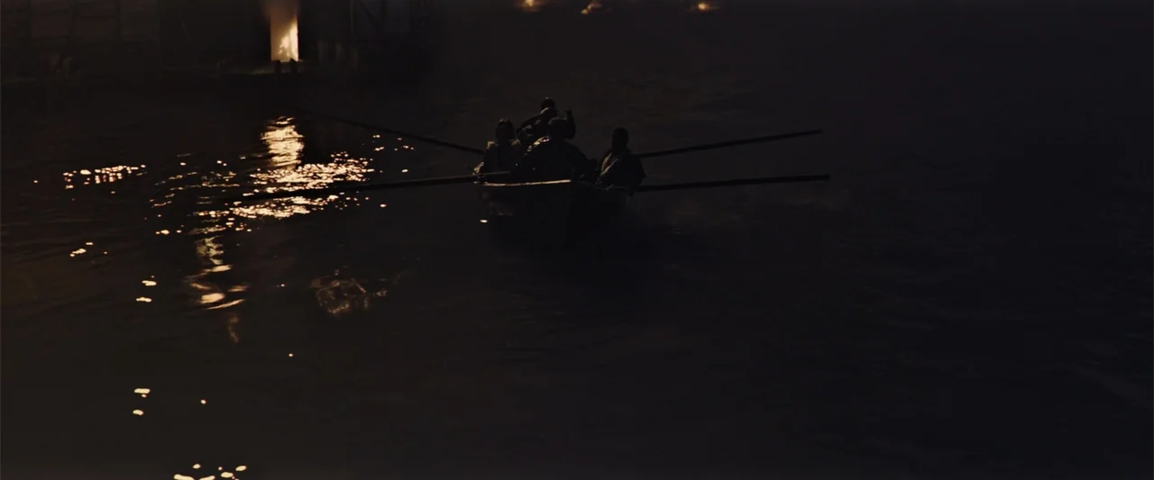 King Arthur: Legend of the Sword (2017) movie still: wide — A dark river at night flows past a dimly lit wooden structure. Four men row a small boat…