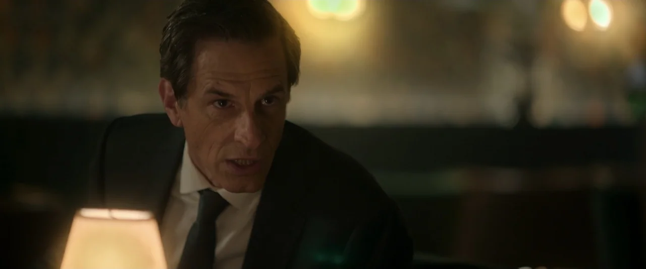 Doublespeak (2021) movie still: medium — A dark restaurant interior features a man sitting at a booth under dim lamps. The man wears a dark suit and…