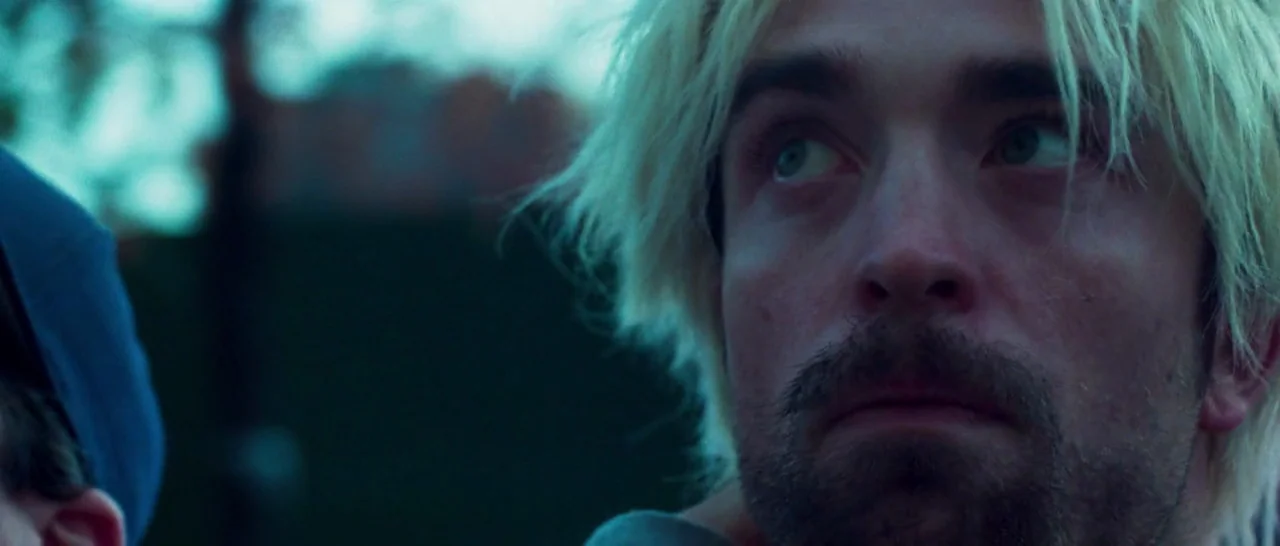Good Time (2017) movie still: closeup — An outdoor urban environment is visible behind a subject in low light conditions. A man with bleach blonde…