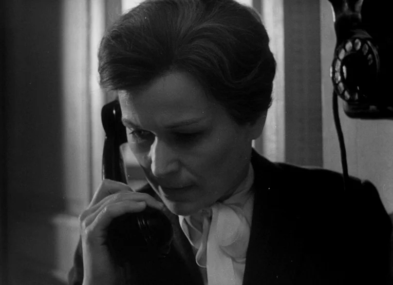 Wings (1966) movie still: closeup — A dark office environment features a woman using a vintage rotary telephone. The woman listens intently to the…